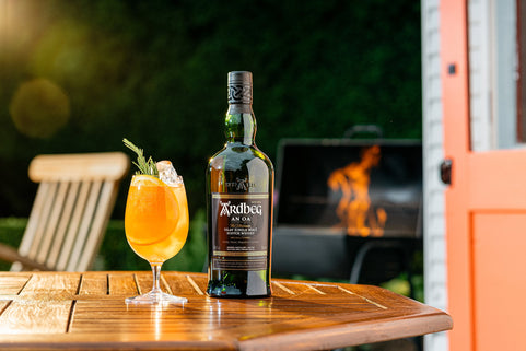 Bottle of Ardbeg An Oa whisky on a wooden table with a cocktail in the foreground, outdoor BBQ setting.